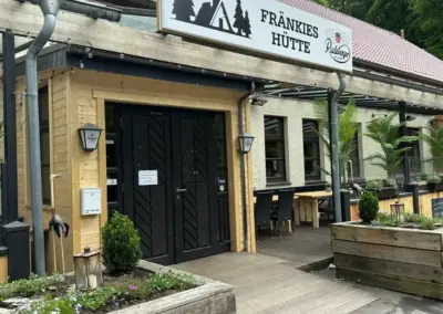 Eingang zu Fränkies Hütte mit Holzverkleidung und Pflanzen im Vordergrund.