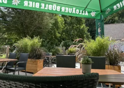Tische und Stühle aus Rattan im Außenbereich unter einem grünen Sonnenschirm.