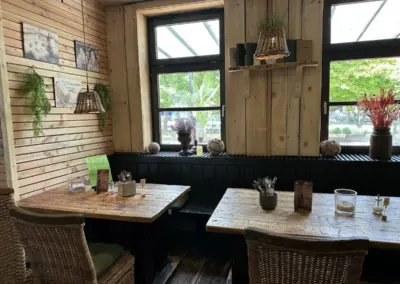 Innenansicht eines Restaurants mit zwei Holztischen, Stühlen und Fenstern mit Pflanzen im Hintergrund.